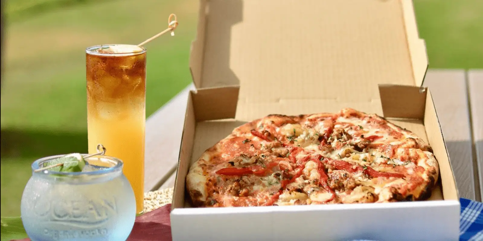 A freshly baked pizza with various toppings is placed in an open pizza box on an outdoor table, perfect for enjoying the best dinner in Kihei. Next to the pizza, there's a tall glass of iced tea with a stir stick and a clear glass bowl containing a beverage garnished with a slice of lime.
