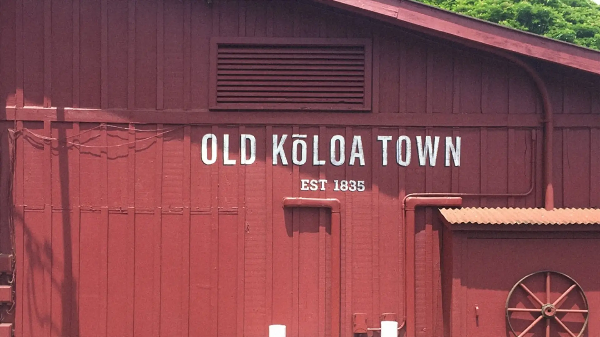 A rustic red building, one of the Best Places to Visit on Kauai, with "Old Kōloa Town" and "Est 1835" painted on its side in white letters. A small window with a horizontal vent is above the sign, and the building boasts visible wooden paneling and an awning over a doorway.