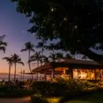 A tropical outdoor restaurant in West Maui is illuminated with warm string lights at dusk. Palm trees and lush greenery surround the venue, creating a cozy and inviting atmosphere. The sky is a gradient of blue and orange as the sun sets in the background, making these restaurants ready for your visit.