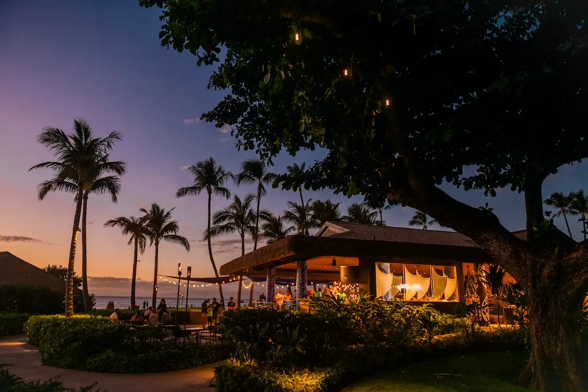 A tropical outdoor restaurant in West Maui is illuminated with warm string lights at dusk. Palm trees and lush greenery surround the venue, creating a cozy and inviting atmosphere. The sky is a gradient of blue and orange as the sun sets in the background, making these restaurants ready for your visit.