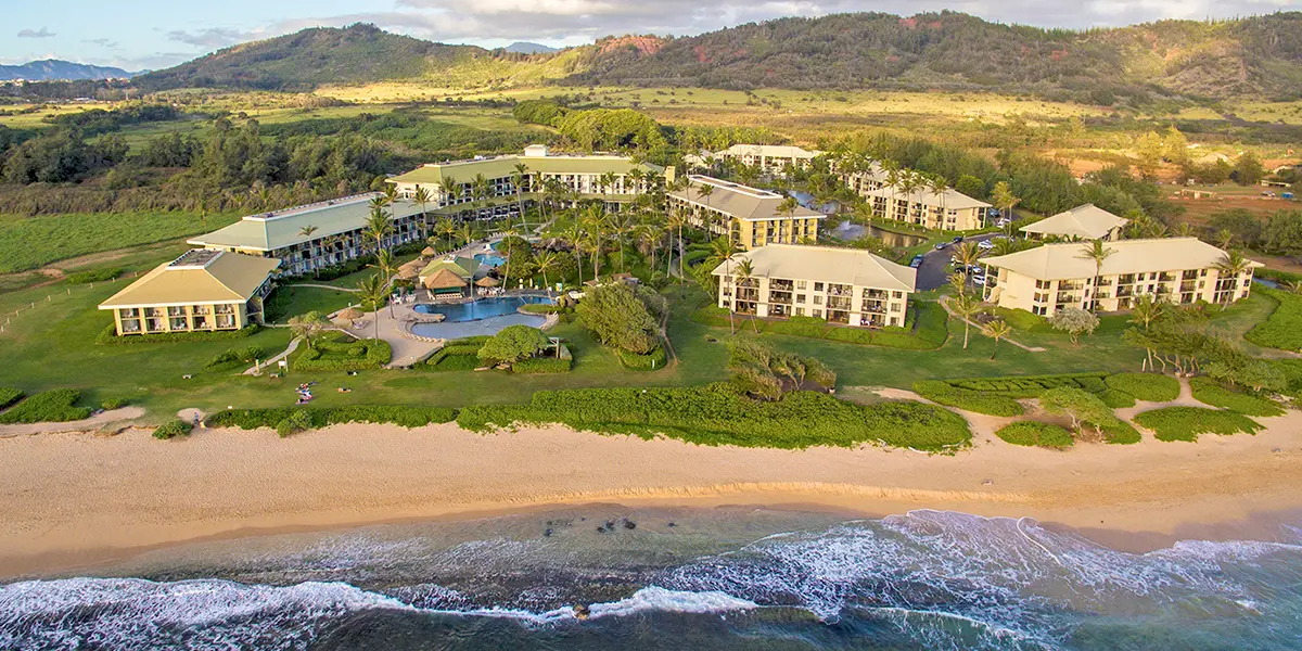 Aerial view of a beachfront resort consisting of multiple buildings with a pool area surrounded by palm trees. The sandy beach leads to the ocean, with gentle waves. Lush greenery and mountainous terrain stretch into the background under a partly cloudy sky.