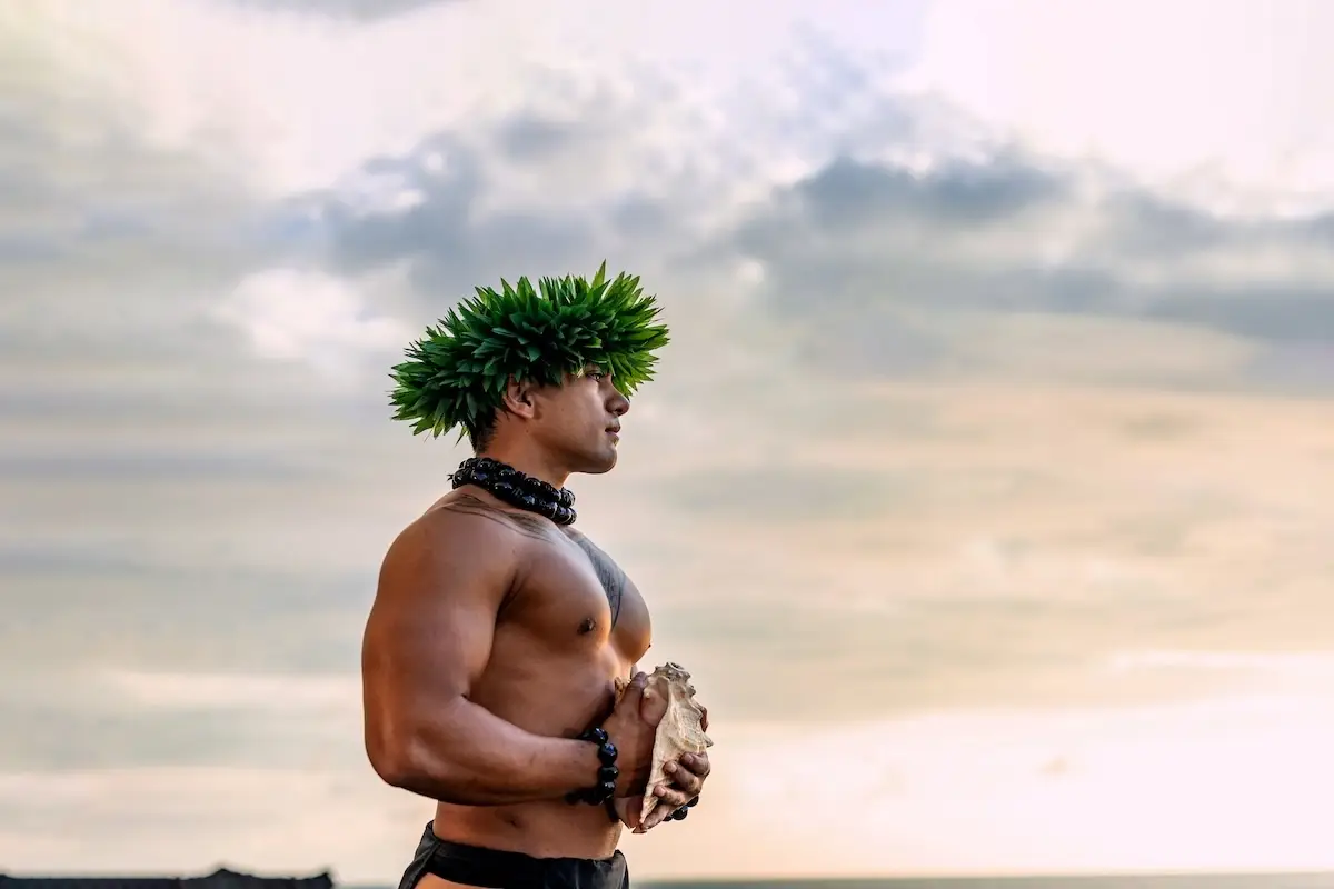 A shirtless man stands outdoors at sunset with the ocean and sky in the background. Engaged in Kona teenager activities, he wears a leafy headpiece and traditional necklace, holding a large shell or conch in his hands.