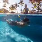 Underwater view of a boy swimming in a pool wearing goggles, with another child standing near the edge of the pool. Bright sunlight and palm trees are visible in the background, creating a tropical atmosphere perfect for Kona teenager activities.