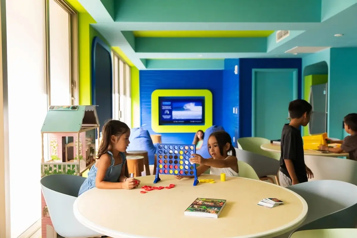Children enjoy various games in a brightly lit room with vibrant green and blue walls typical of Waikiki family activities. Two girls play Connect Four at a round table in the foreground, while other kids engage in different activities at tables in the background. A TV is mounted on the wall.