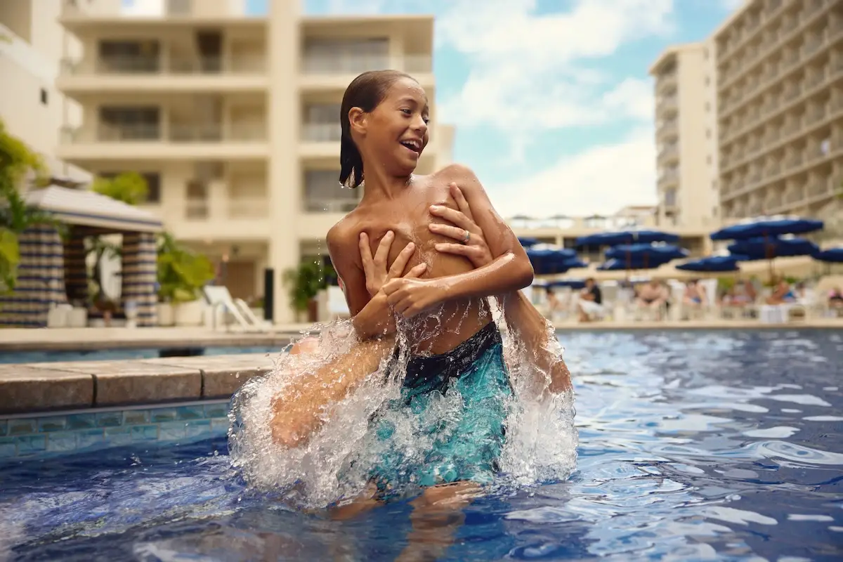 Two boys are playing in a pool. One boy is holding another boy in a playful manner, both laughing and having fun. Apartment buildings and blue umbrellas in lounge areas are in the background. The sky is blue with a few clouds, perfect for enjoying Waikiki family activities together.