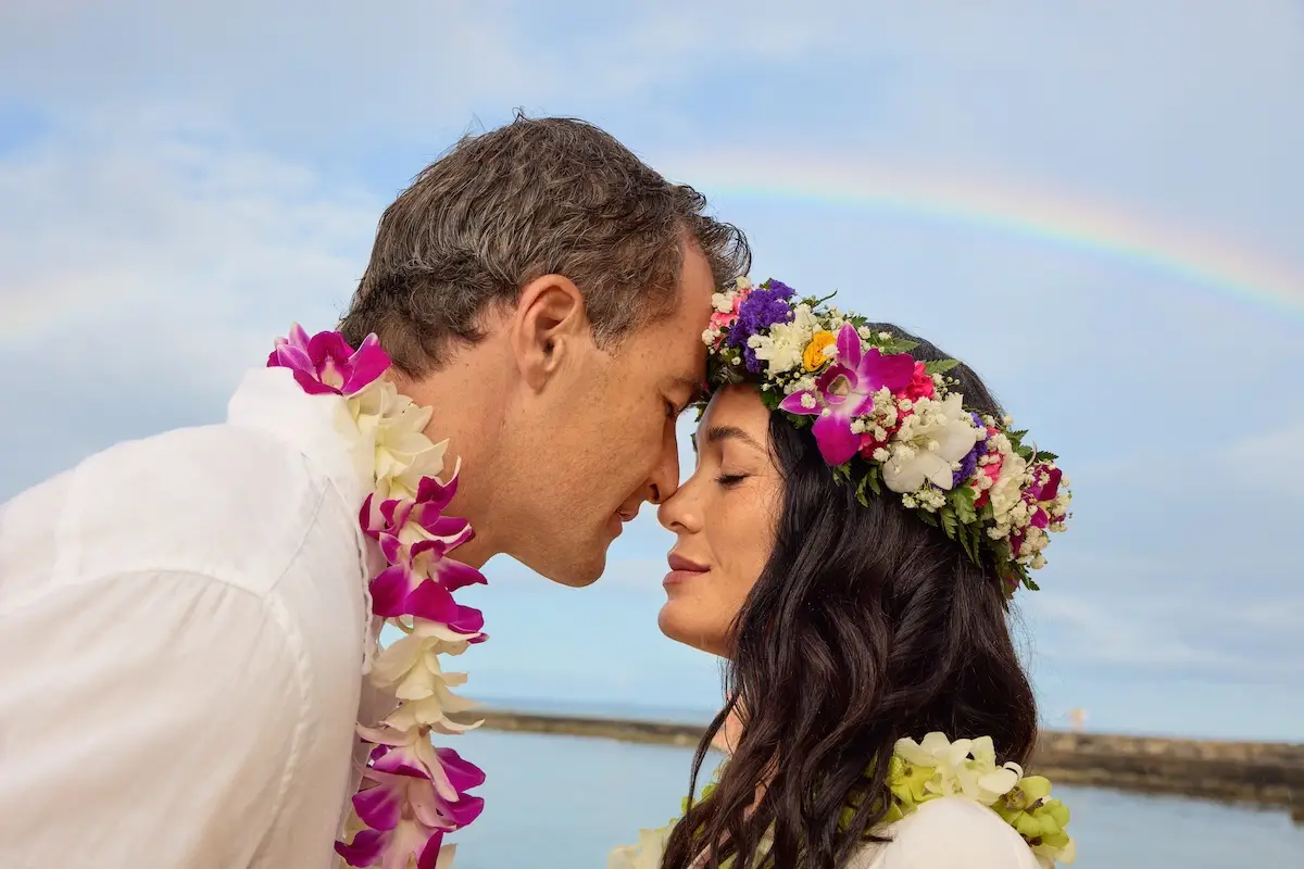 A couple wearing floral leis and headpieces leans in close, foreheads touching tenderly, with a serene ocean and a rainbow in the background. Both wear white attire and appear to be enjoying a romantic moment outdoors—a perfect snapshot of Waikiki for couples seeking tranquility.