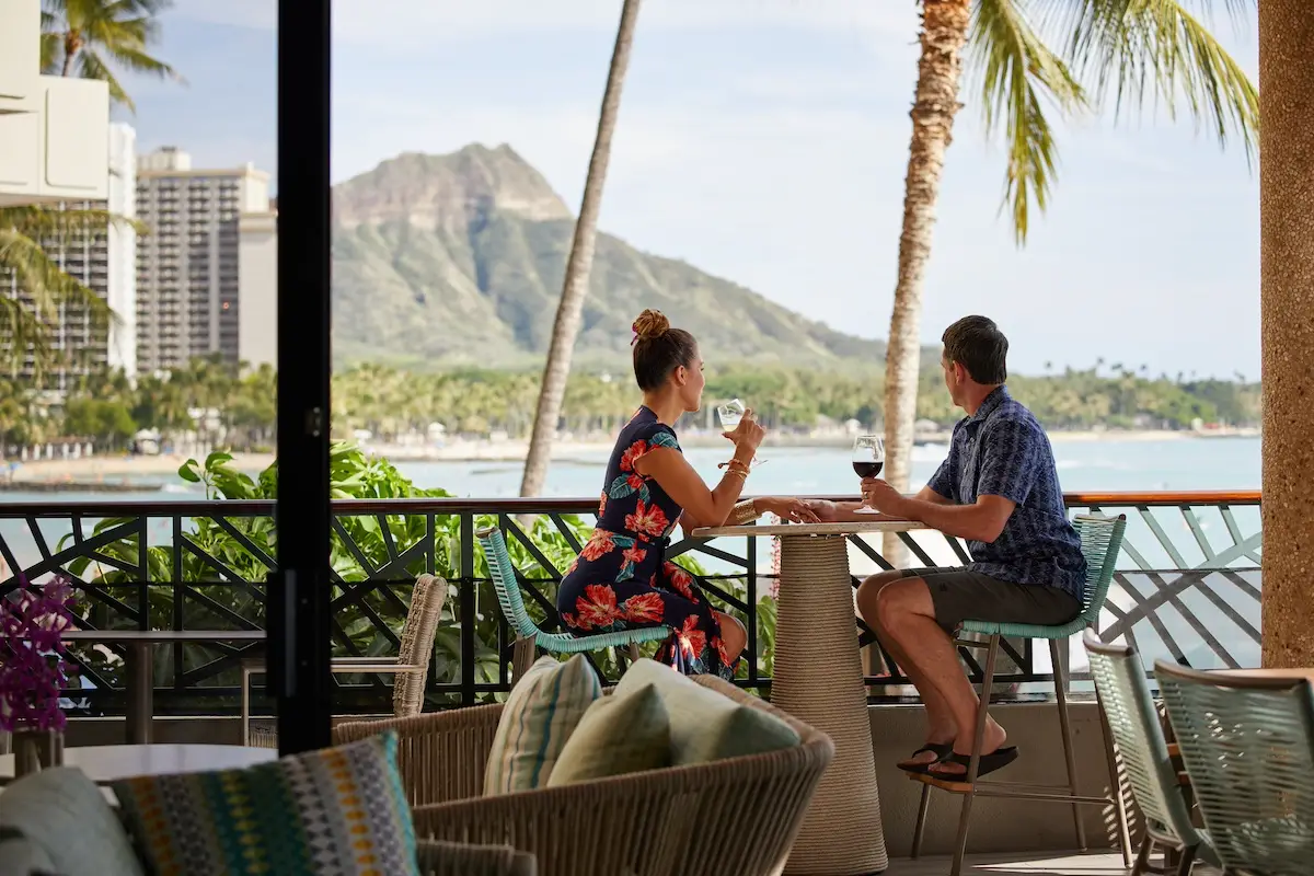 A woman in a floral dress and a man in a blue shirt sit at a high table on a balcony, enjoying drinks. They overlook the scenic Waikiki beach, palm trees, and a mountain under a clear sky. It's an idyllic setting for couples with cozy outdoor furniture in the foreground.
