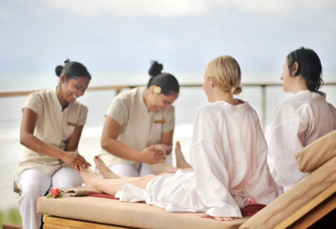Four individuals are seated on lounge chairs, three wearing white uniforms providing foot massages to a person dressed in a white robe. The serene outdoor setting, complete with a soft-focus sky and distant landscape, evokes the tranquility of Valentines Day Gifts from Hawai'i.