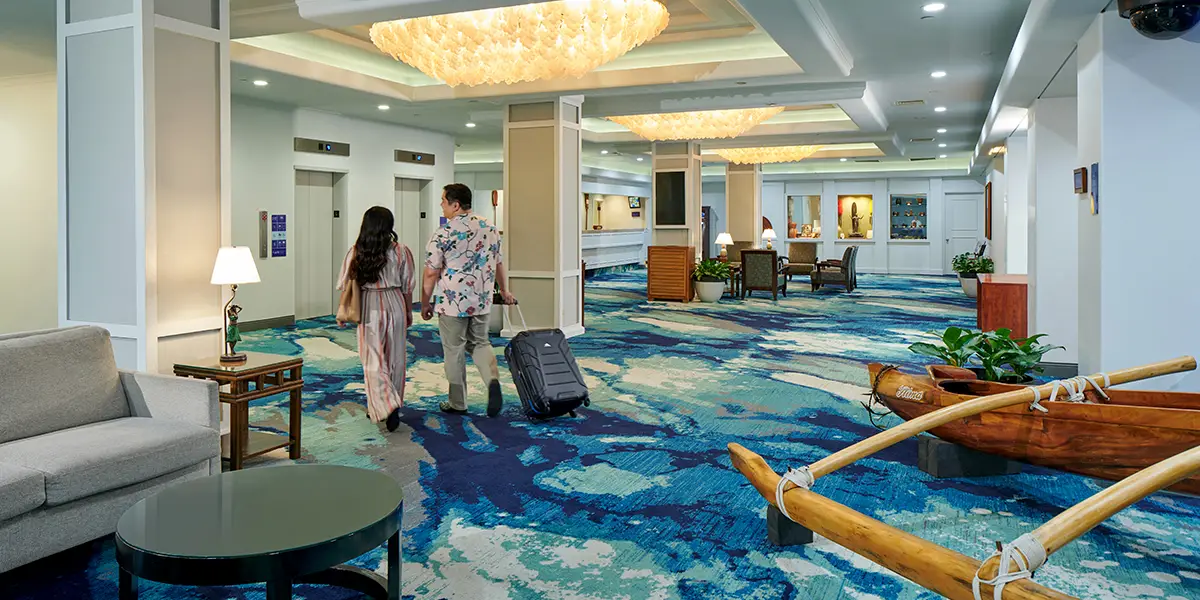 A couple walks through a spacious, brightly-lit lobby at Ohana Waikiki East by Outrigger, featuring a blue patterned carpet. The man pulls a suitcase on wheels while the woman walks beside him. They head toward the modern reception area adorned with a decorative wooden canoe.