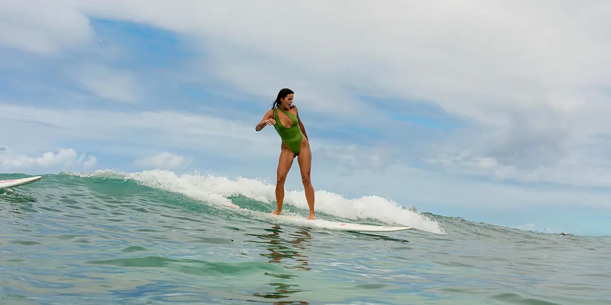 A person wearing a green swimsuit surfing on a small wave under a cloudy sky near Ohana Waikiki East by Outrigger. The ocean appears calm with gentle waves, and the surfer maintains balance as they ride the wave.