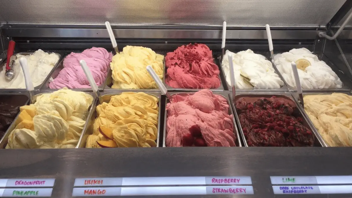 A display case showcasing the best desserts of Maui Wailea features various flavors of gelato in metal containers. Flavors include dragonfruit, pineapple, lilikoi, mango, raspberry, strawberry, raspberry sorbet, lime, and dark chocolate raspberry. Scoops are visible among the bright, colorful gelato.