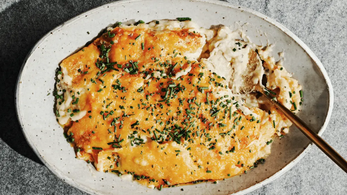 An oval ceramic dish filled with a cheesy mashed potato casserole, topped with melted golden cheese and sprinkled with chopped chives. A spoon is partially dipped into the casserole. This delectable addition to potato dishes is placed on a gray surface.
