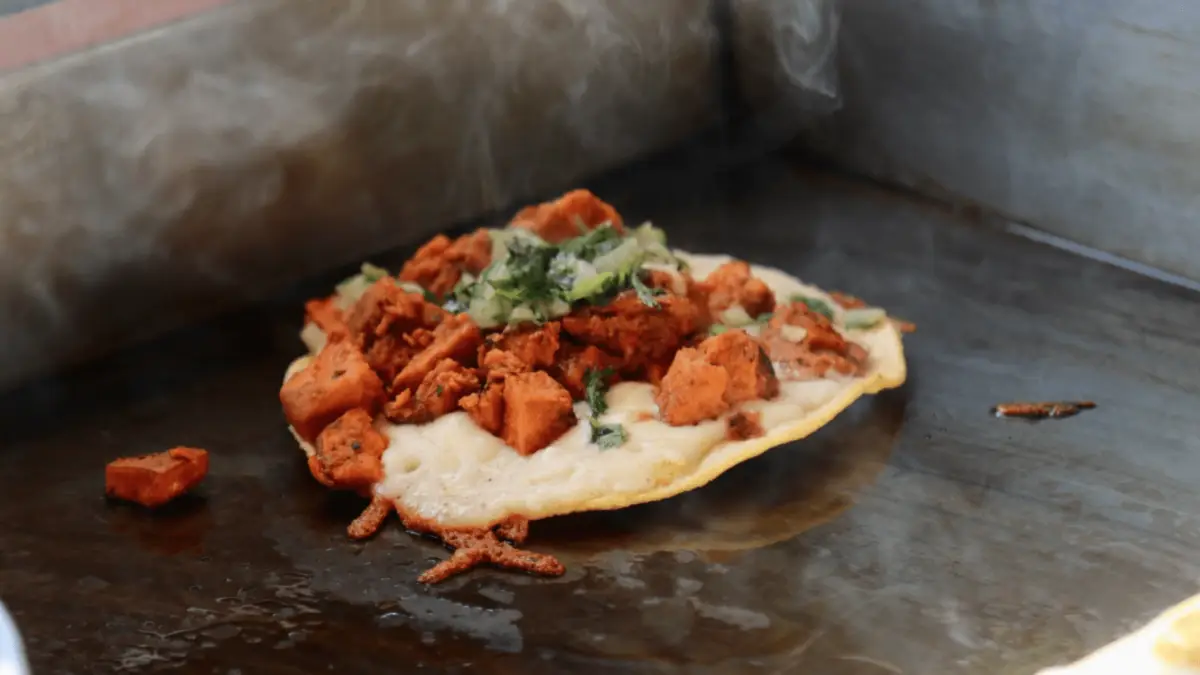 A sizzling taco with a crispy tortilla is being cooked on a hot griddle. It is topped with small chunks of seasoned and cooked meat, garnished with fresh herbs and onions. Steam rises from the taco as it cooks, reminiscent of hearty potato dishes that bring warmth and comfort.