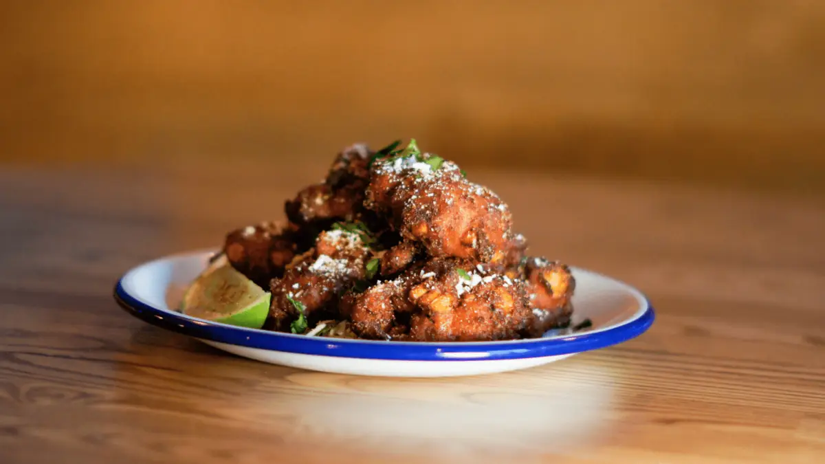 A white plate with a blue rim holds a stack of crispy, golden-brown fried chicken wings topped with grated cheese and fresh herbs. A slice of lime rests beside the wings on a wooden table adorned with assorted potato dishes, set against a warm, blurred background.