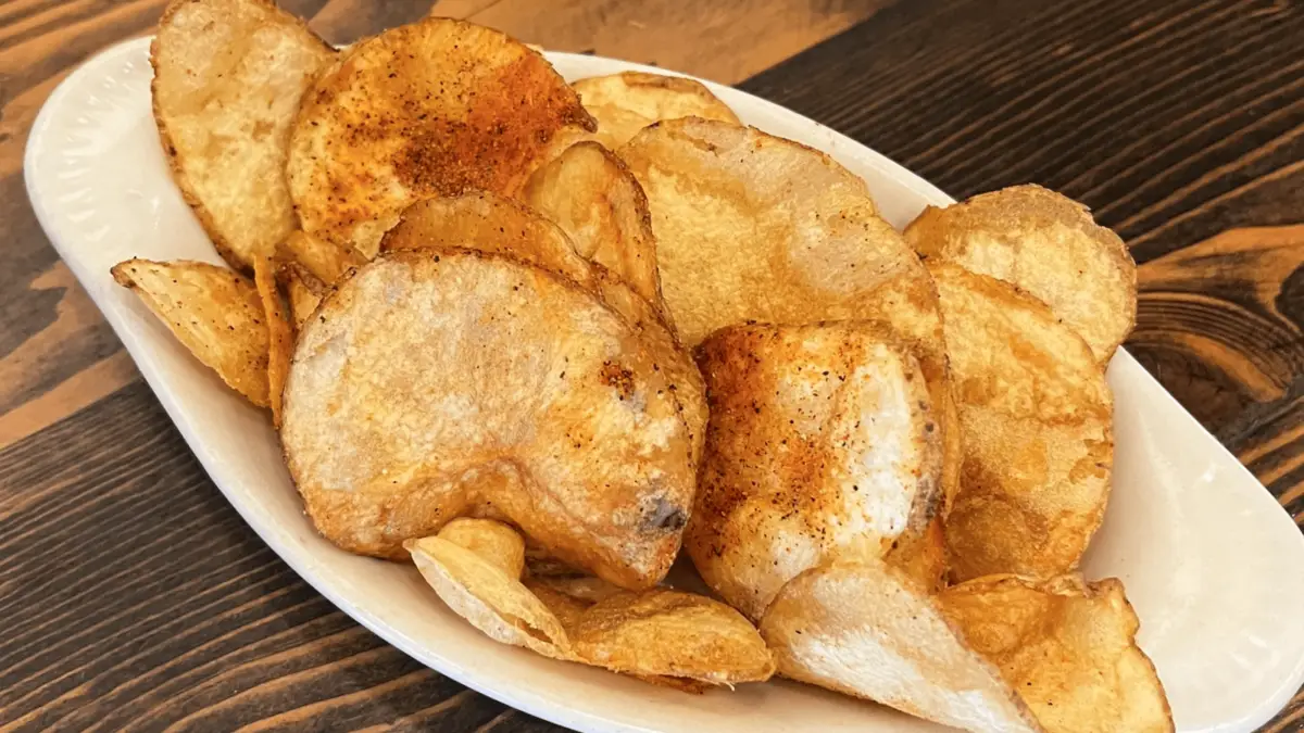 A white oval plate filled with crispy, golden potato chips sprinkled with seasoning. The chips are arranged in a semi-upright position, showcasing one of the finest potato dishes, and the plate is placed on a wooden table.