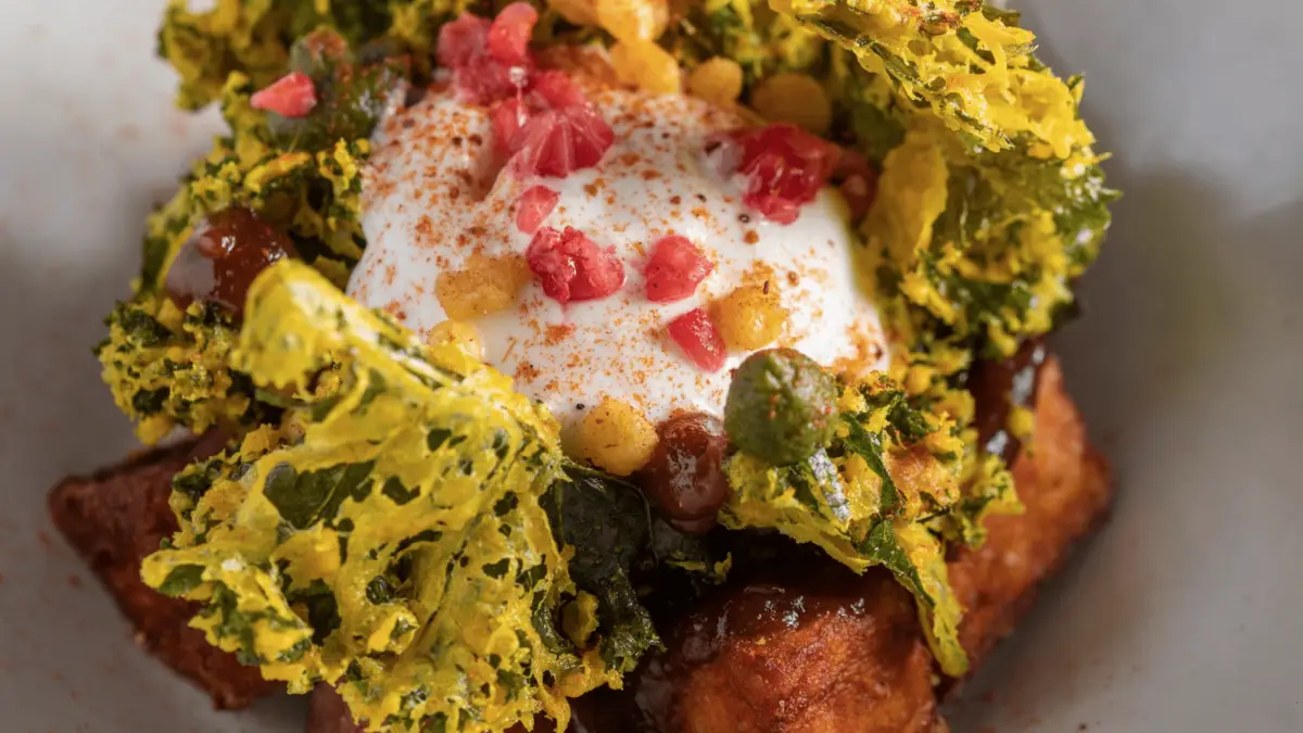 A close-up of a gourmet dish perfect for bay area brunch, featuring a perfectly poached egg topped with pink bits, dusted with spices, surrounded by crispy spinach leaves, and garnished with small yellow spheres. The ensemble is placed on a layer of seared meat chunks.