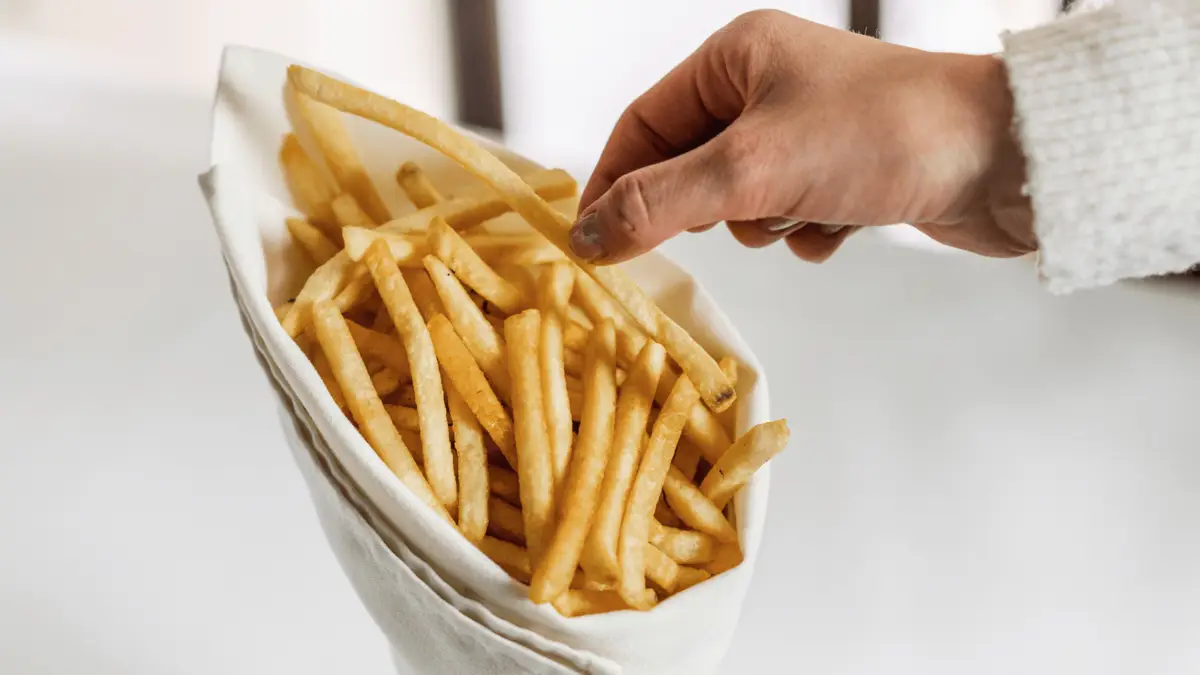 A person is reaching for a French fry from a white cloth basket, filled with a generous portion of golden, crispy potato dishes. The hand is slightly above the fries, with fingers gently grasping one fry. The background is blurred and white.