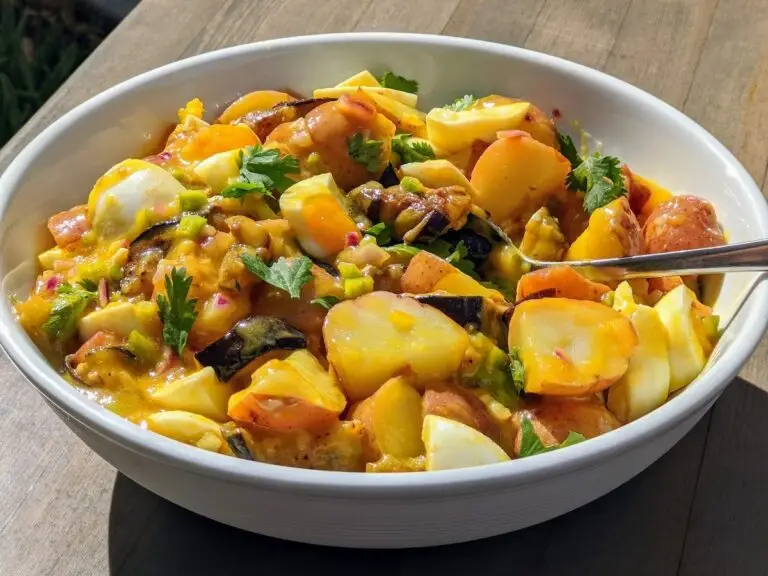 A white bowl filled with colorful potato salad is on a wooden table, perfect for a Memorial Day BBQ. The salad includes yellow potatoes, hard-boiled eggs, chopped vegetables, and fresh herbs with a creamy dressing. A metal spoon rests in the bowl as sunlight enhances the vibrant colors of the dish.