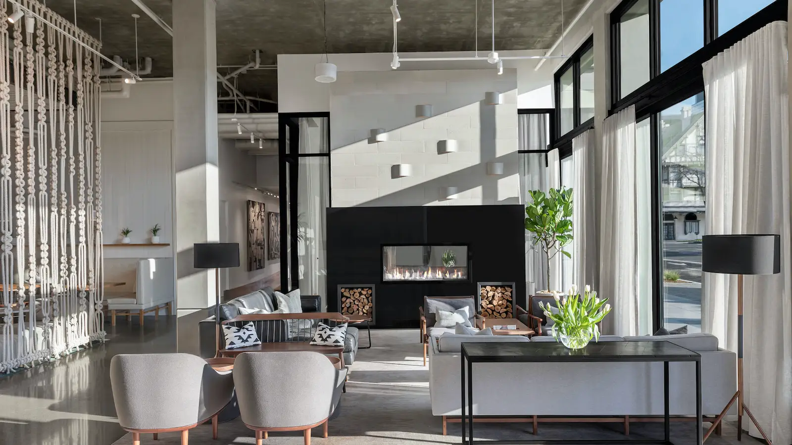 A modern lounge area with stylish furniture including gray chairs and wooden coffee tables. A sleek, black fireplace is centrally placed on a white wall. Natural light streams through large windows with white curtains. Various plants add greenery to the space, embodying a vibe found in the best South Bay hotels.