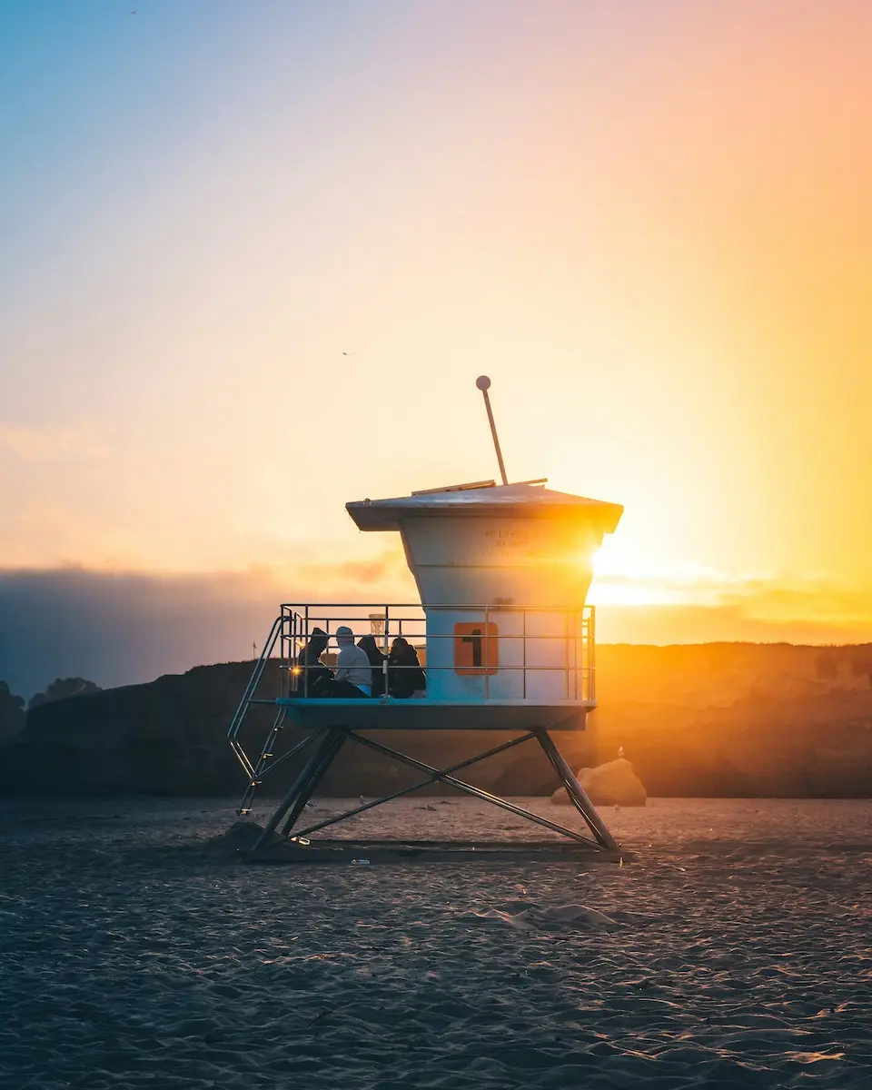 A lifeguard tower on the beach is bathed in warm sunlight at sunset. Two people sit on the platform, facing the ocean along Central Coast California. The sky displays a gradient from deep blue to orange, with a hint of clouds in the background.