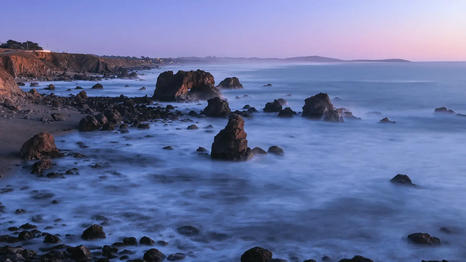 A coastal scene at dusk in Sonoma Wine Country features large, jagged rocks emerging from calm, misty waters. The sky has a gradient from light pink to deep blue. The rocky shoreline and cliffs extend into the distance, with a serene, ethereal atmosphere created by the soft lighting—perfect for summer adventures.