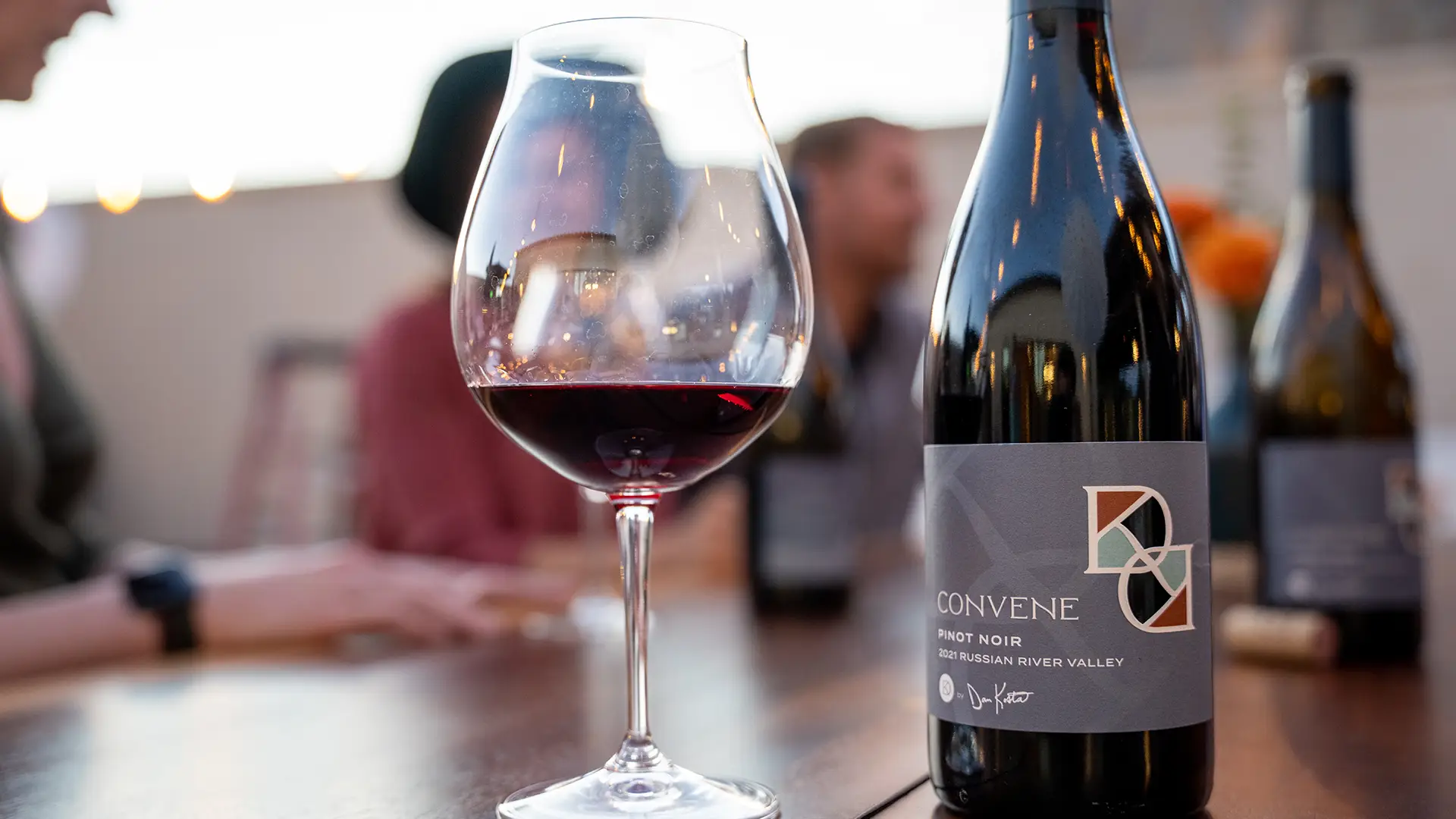 A close-up of a glass of red wine next to a bottle labeled "Convene Pinot Noir - Russian River Valley" on a wooden table. In the background, reminiscent of Sonoma tasting rooms, three people seated around the table are blurred, engaging in conversation.
