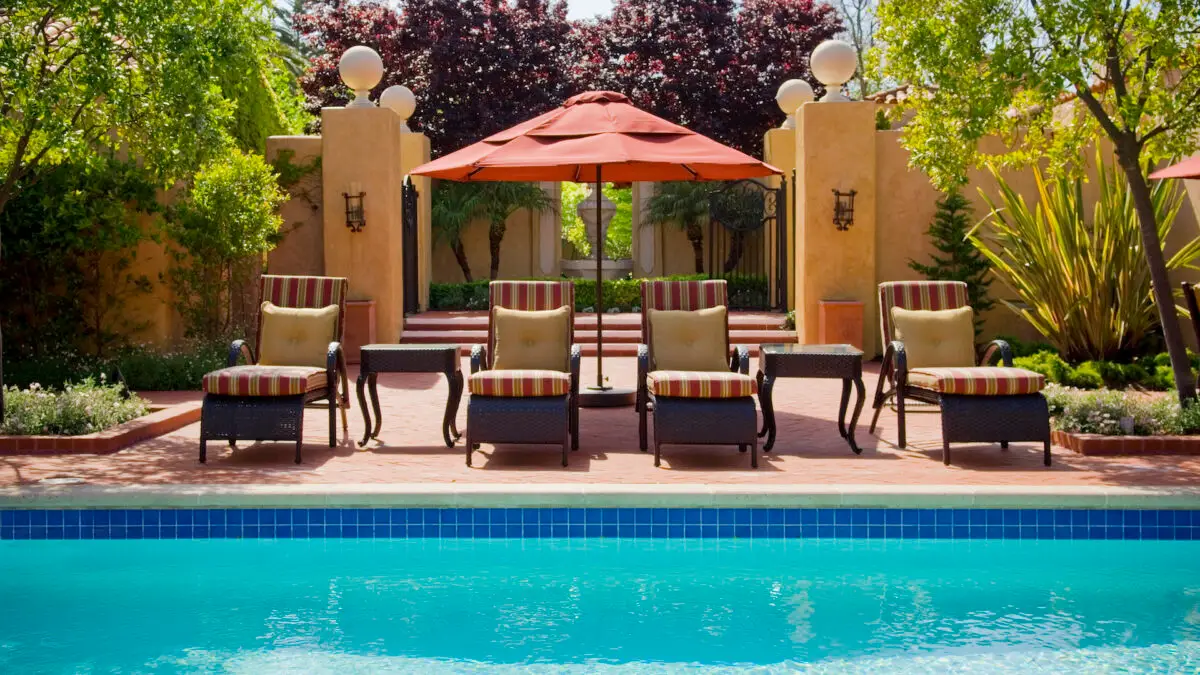 A serene outdoor pool area at one of the best romantic hotels in Monterey features five cushioned lounge chairs with striped pillows and matching side tables, all shaded by a large red umbrella. The scene is framed by vibrant greenery and a stone wall with trees and bushes in the background.