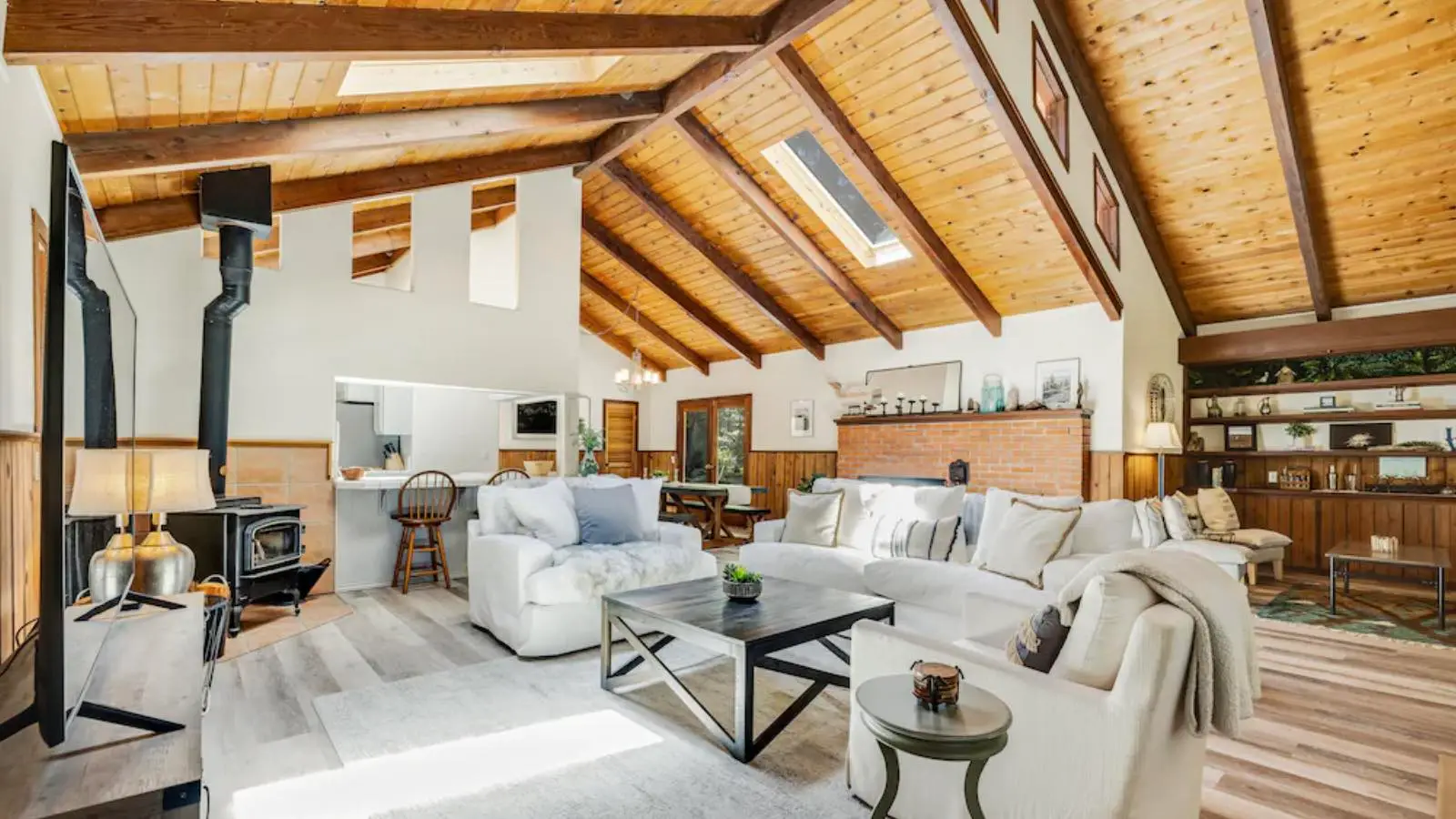 A spacious living room, ideal for Bay Area Vacation Rentals, features high, sloped wooden ceilings with exposed beams and skylights. Furnished with white sofas, a coffee table, and a wood-burning stove, the room opens into a bright dining area. Various décor accents and bookshelves add warmth to the space.