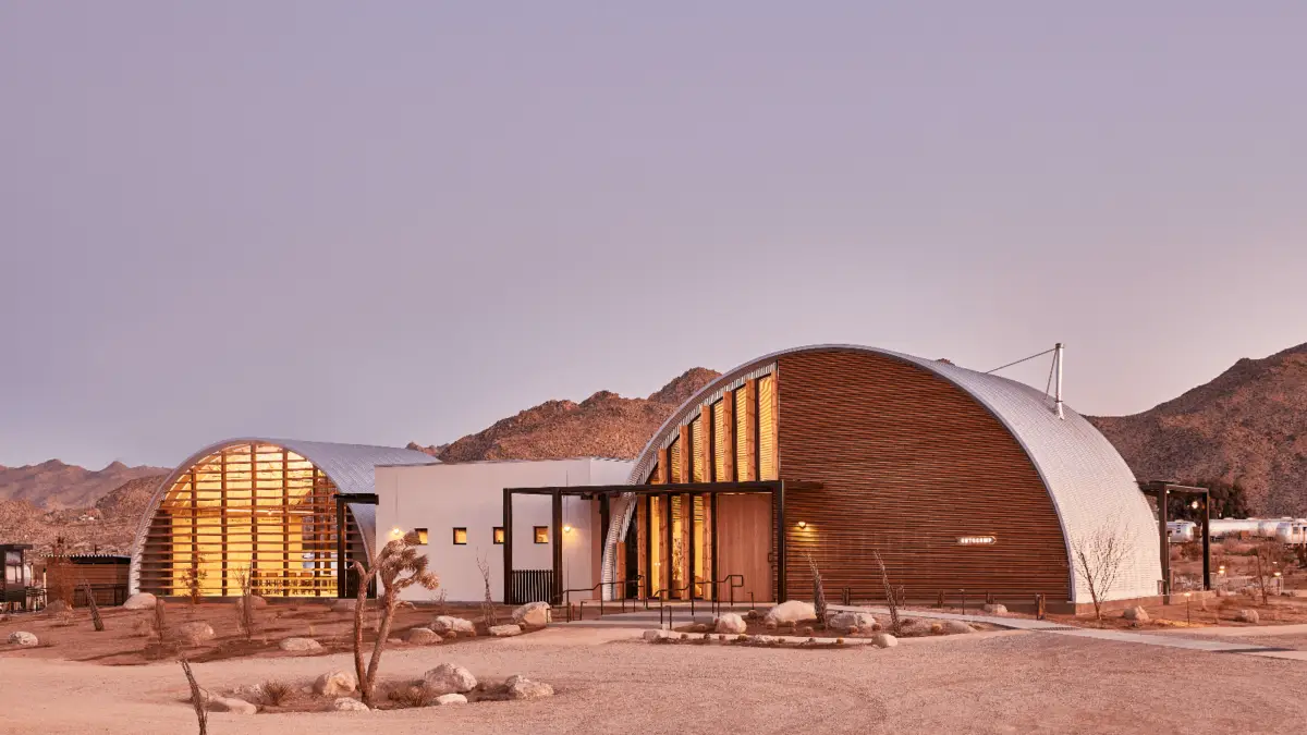 A modern, arched building with large windows is situated in a desert landscape at dusk. The structure is illuminated from within, showcasing a warm glow. This architecturally iconic example of California hotels is surrounded by sparse vegetation and rocky terrain, with mountains visible in the background.