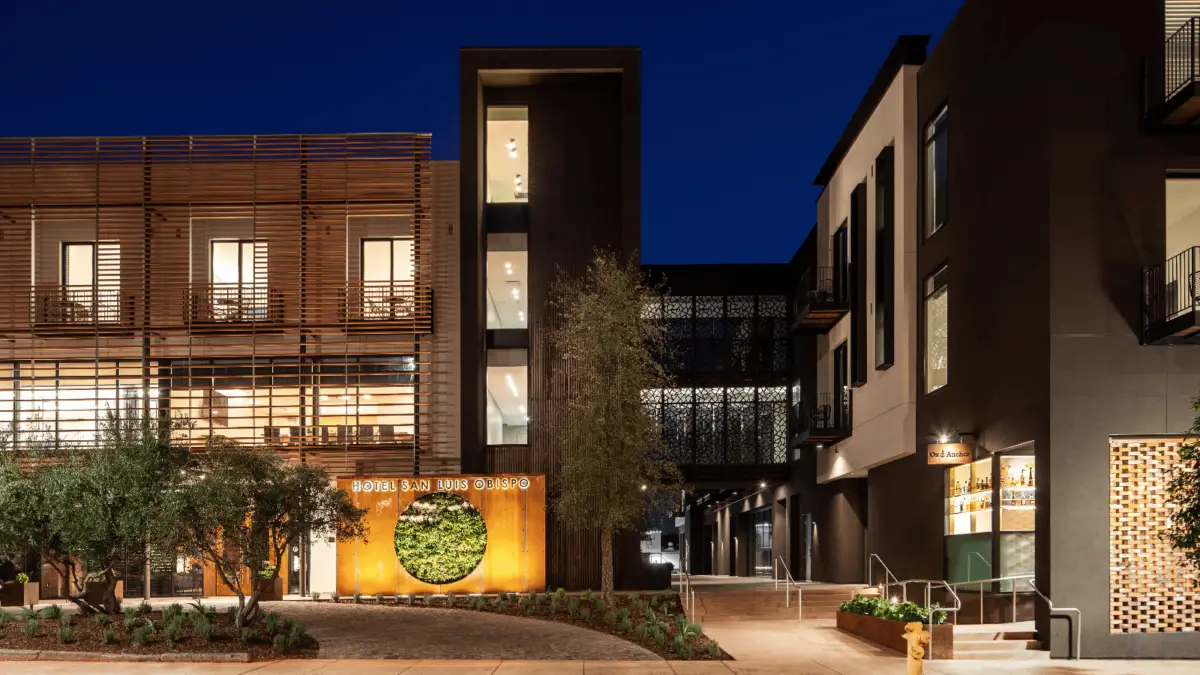 A well-lit hotel entrance at night, featuring modern architecture with wooden slats and metal elements, embodies the charm of Architecturally Iconic California Hotels. The sign reads "Hotel San Luis Obispo," surrounded by greenery. Large windows adorn both visible buildings, with steps and a pathway leading to the entrance.