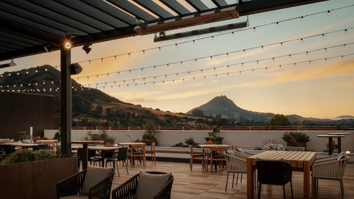 Rooftop patio dining area at sunset, with string lights overhead. Tables and chairs are arranged around the space, providing a view of distant mountain peaks and a brightly colored sky transitioning from blue to orange hues. Lush greenery surrounds the seating, reminiscent of central California missions.