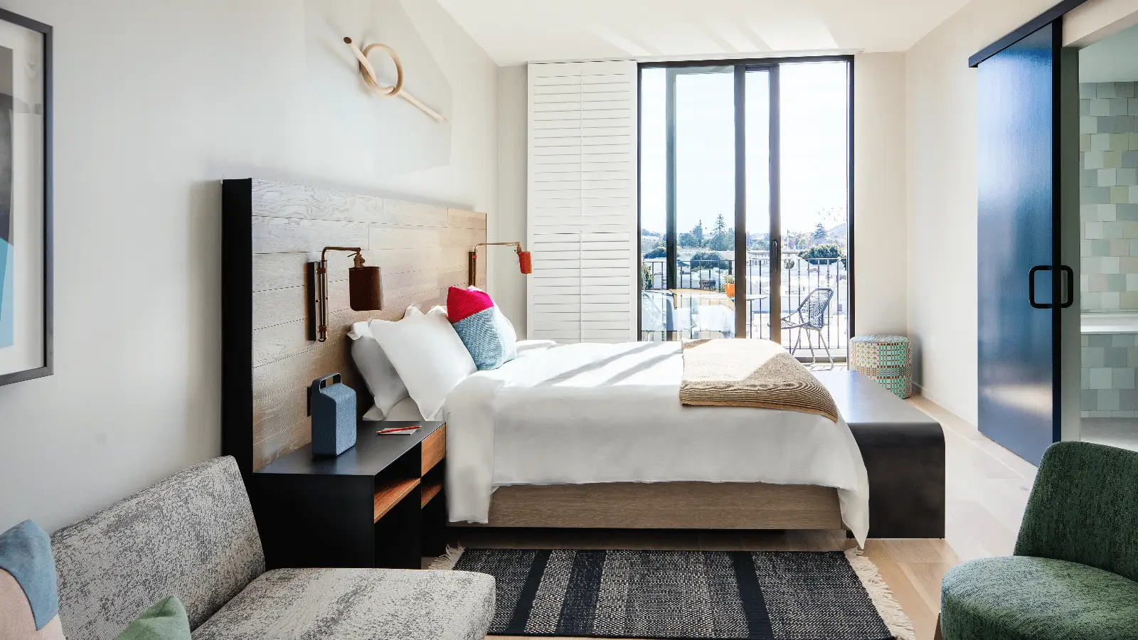 A modern hotel room in one of the architecturally iconic California hotels, featuring a large bed with white linens and colorful pillows. Nightstands with lamps flank the headboard. The room includes a sofa, a rug, and a balcony view with chairs outside large floor-to-ceiling windows.