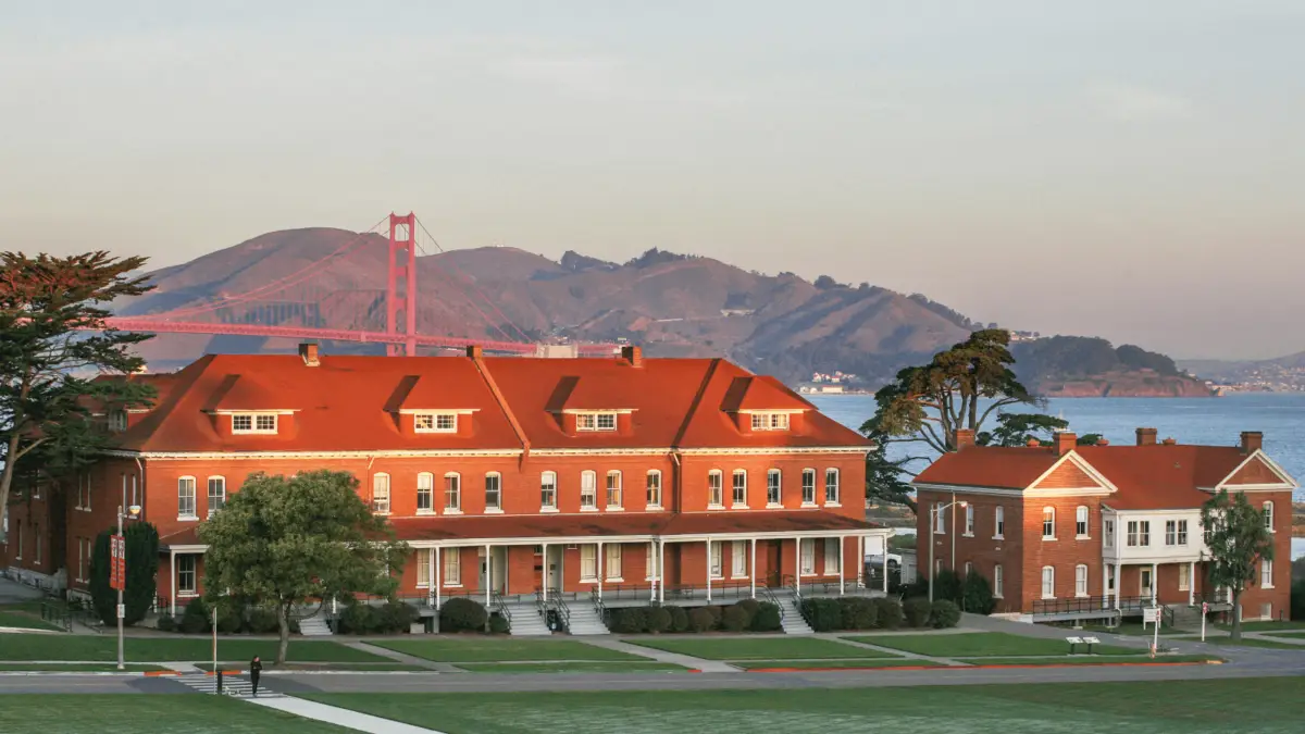 A scenic view of red-roofed brick buildings with white trim, surrounded by lush green lawns and trees. In the background, the architecturally iconic Golden Gate Bridge stretches across the bay with rolling hills under a clear, pastel-colored sky at sunset.
