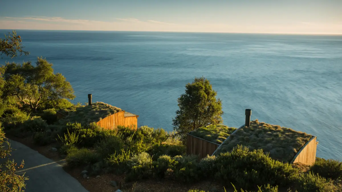Two wooden cabins with green rooftops blend into the lush vegetation on a hillside overlooking a calm, expansive ocean under a clear sky. The scene, reminiscent of Big Sur secrets, is peaceful and serene with an emphasis on nature and tranquility.