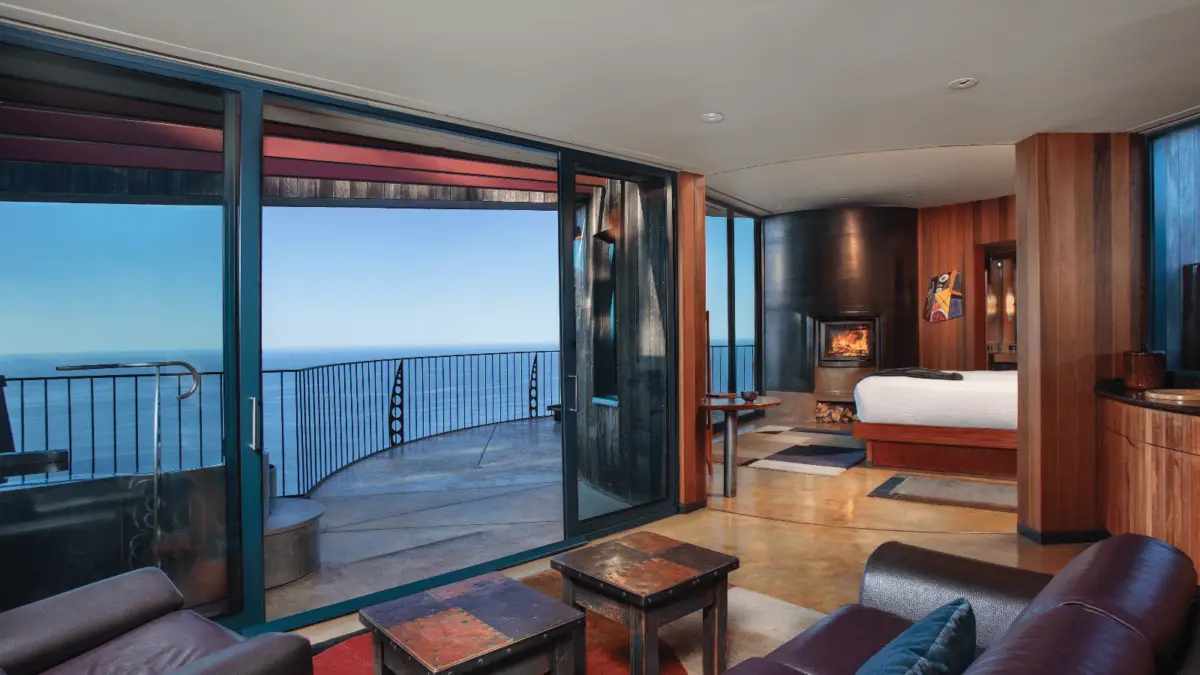 A modern, open-concept living area with glass sliding doors leading to a balcony overlooking the ocean. The room is furnished with a leather sofa, a fireplace, wooden accents, and a bedroom area. Natural light floods the space, highlighting the contemporary decor reminiscent of architecturally iconic California hotels.