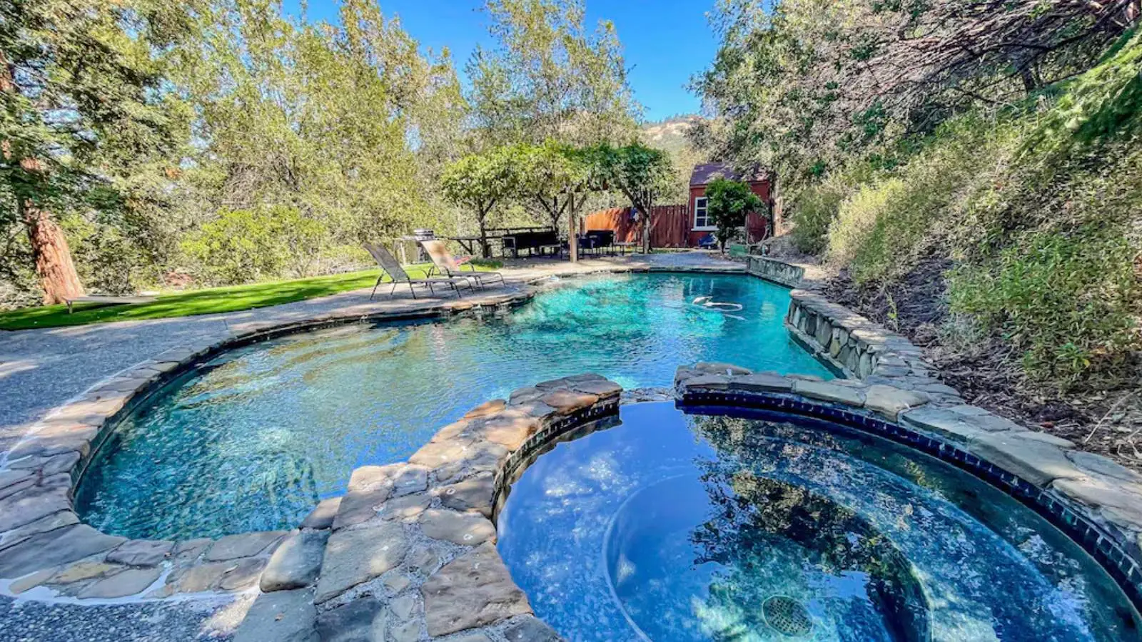 A tranquil outdoor pool with an attached hot tub surrounded by a stone deck. Two lounge chairs sit on the grassy lawn next to the pool. Trees and foliage encircle the area, providing shade and privacy. A picnic table and a red shed are visible in the background, making it ideal for Bay Area Vacation Rentals.
