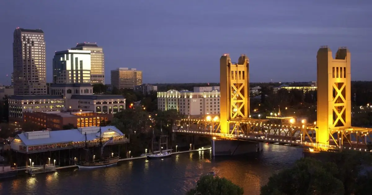 A nighttime view of Sacramento's skyline featuring illuminated buildings and the iconic Tower Bridge with its golden-yellow towers reflecting on the Sacramento River. The cityscape includes a mix of modern and historic architecture under a dusky sky.