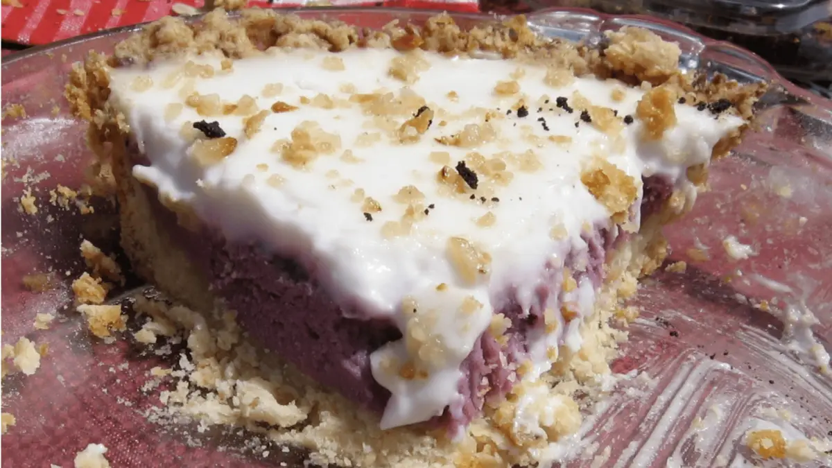 A slice of pie with a crumbly crust, a thick purple berry filling, and a generous layer of white frosting on top. Crumbs are scattered around the slice on a clear glass plate, and the pie appears to have been partially eaten—truly one of the best desserts Maui Wailea has to offer.