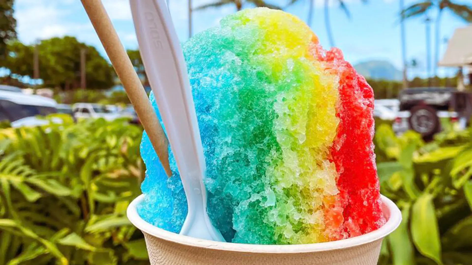 A colorful shaved ice dessert with red, yellow, green, and blue sections is in a white paper cup. A white plastic spoon and a brown paper straw are sticking out of the ice. The background shows lush greenery, palm trees, a blue sky, and parked cars—truly the best shave ice in Poipu.