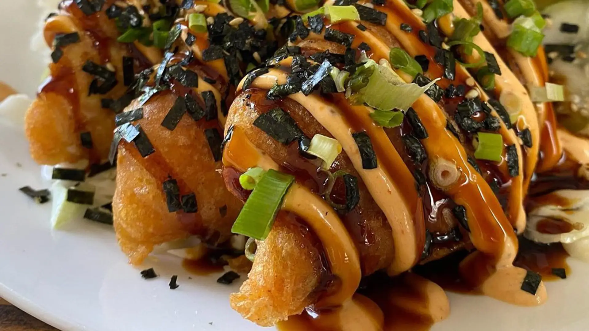 A close-up of crispy tempura sushi rolls drizzled with spicy mayo and eel sauce, garnished with chopped green onions and strips of nori. The rolls are arranged on a white plate, displaying vibrant colors and textures—truly the best lunch Kauai has to offer.