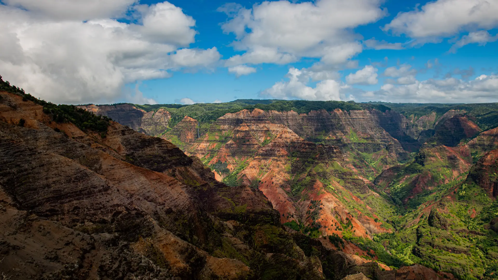 A stunning landscape of Waimea Canyon in Hawaii, one of the best places to visit on Kauai, featuring rugged, colorful canyon walls in shades of red, green, and brown. Lush greenery can be seen along the canyon floors. The sky above is bright blue with scattered white clouds.