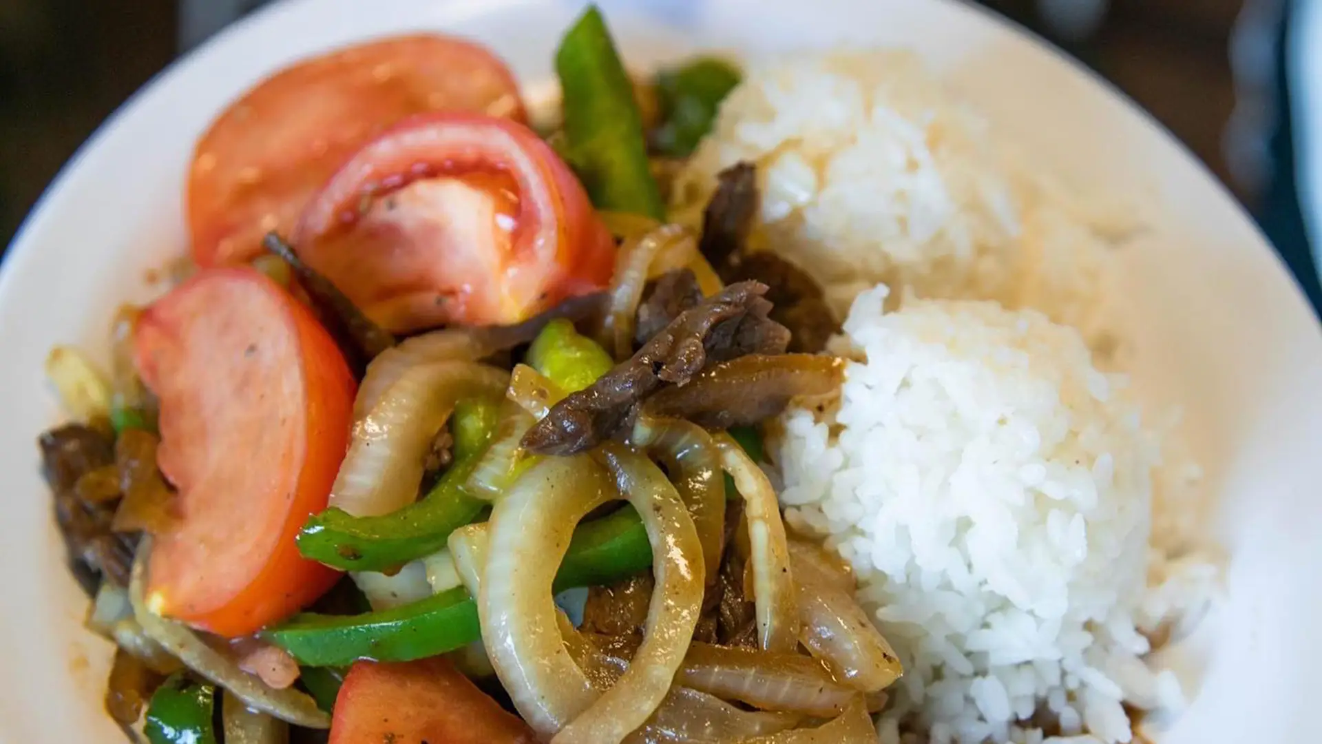 A plate of food featuring stir-fried beef with onions, bell peppers, and tomato slices, accompanied by two portions of white rice. The dish is colorful with a combination of green, red, and brown tones – truly the best lunch Kauai has to offer.