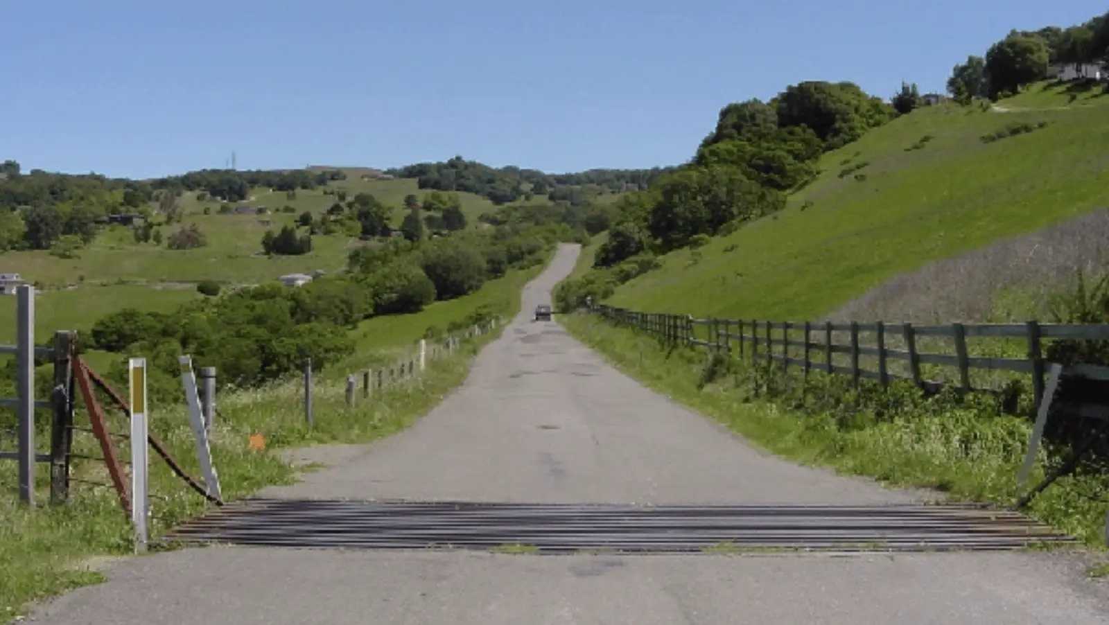 A long, empty road stretches into the distance, surrounded by green fields and hills under a clear blue sky. A cattle guard is visible in the foreground, and a solitary vehicle from Family Friendly Adventures Sonoma County is seen driving away in the distance.