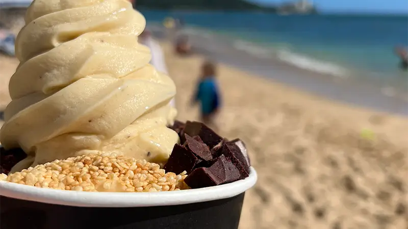 Close-up of a cup of ice cream topped with sesame seeds and chocolate chunks. The background features the sandy shores of Why Waikiki, with blurred figures and blue ocean waves, suggesting a warm, sunny day by the seaside.