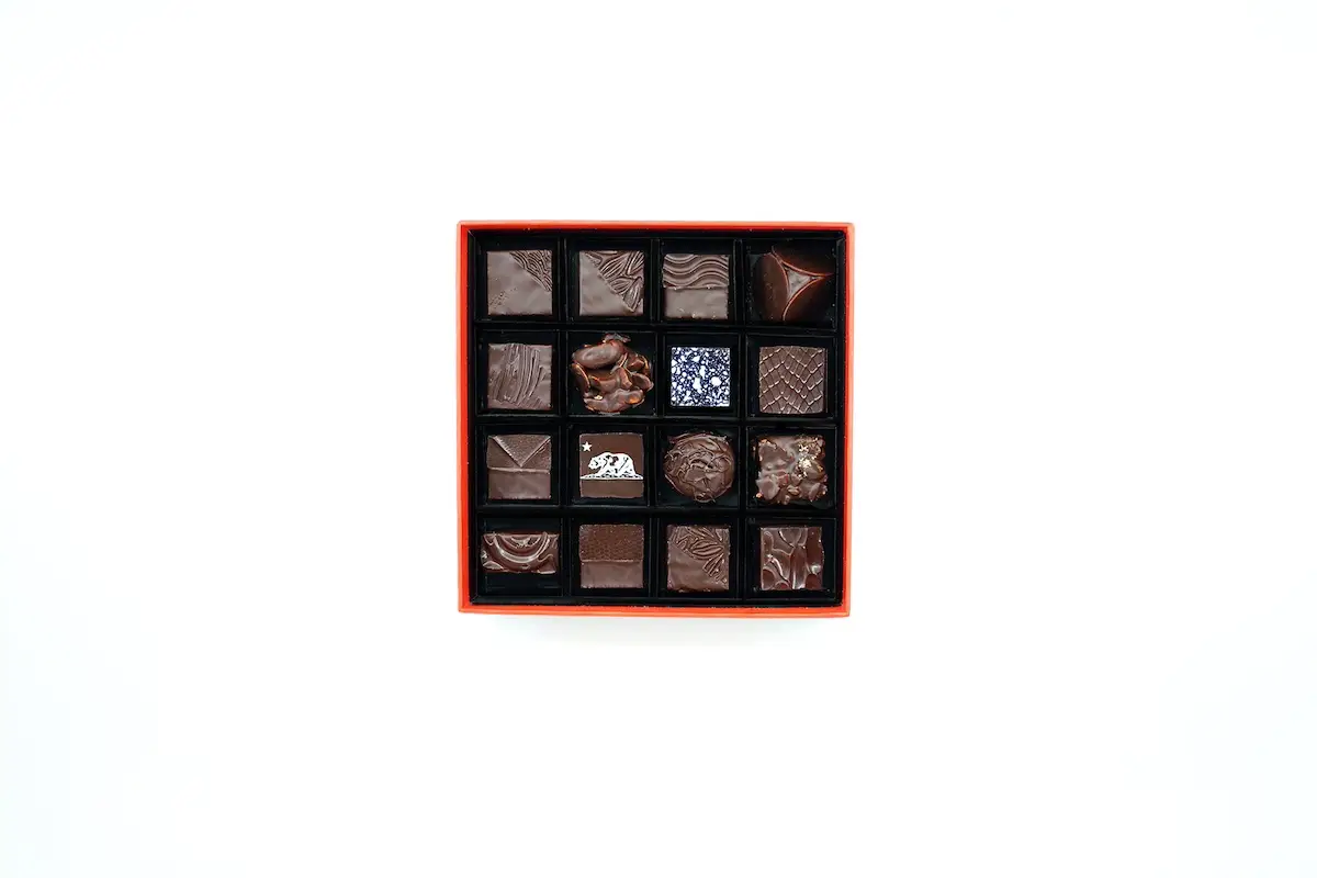 A box of assorted chocolates arranged in a grid with four rows and four columns. Each chocolate, showcasing the health benefits of dark chocolate, has a unique design or topping, varying in shapes and patterns. The box is rectangular with a red exterior and black interior. The background is plain white.