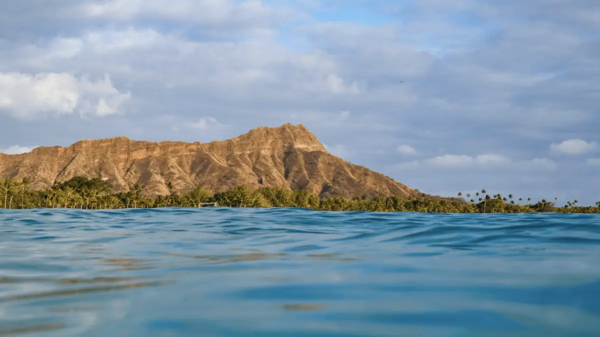 A scenic view of Diamond Head, a volcanic crater in Hawaii, seen from the ocean. The foreground features blue, slightly wavy water ideal for scuba diving, while the background showcases the rugged, brown formations of the crater against a partially cloudy sky. Palm trees line the distant shore.