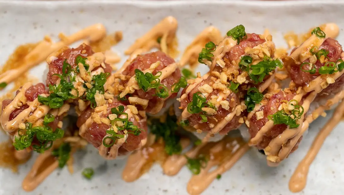 A close-up of a sushi roll topped with diced raw fish, chopped green onions, and crispy tempura flakes, drizzled with orange-colored sauce, arranged on a rectangular white plate.