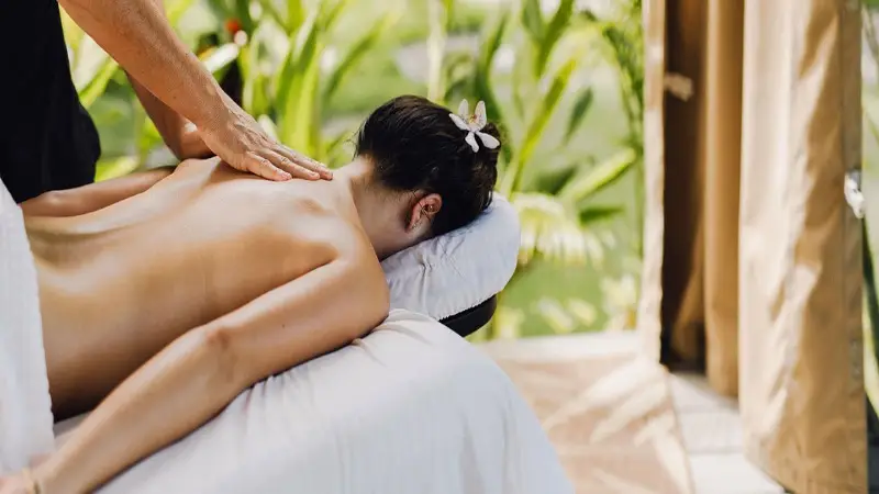 A person is lying face down on a massage table outdoors at the best spa hotel in Maui, with lush greenery in the background. They are receiving a back massage from a therapist. The person has a flower in their hair and appears relaxed.