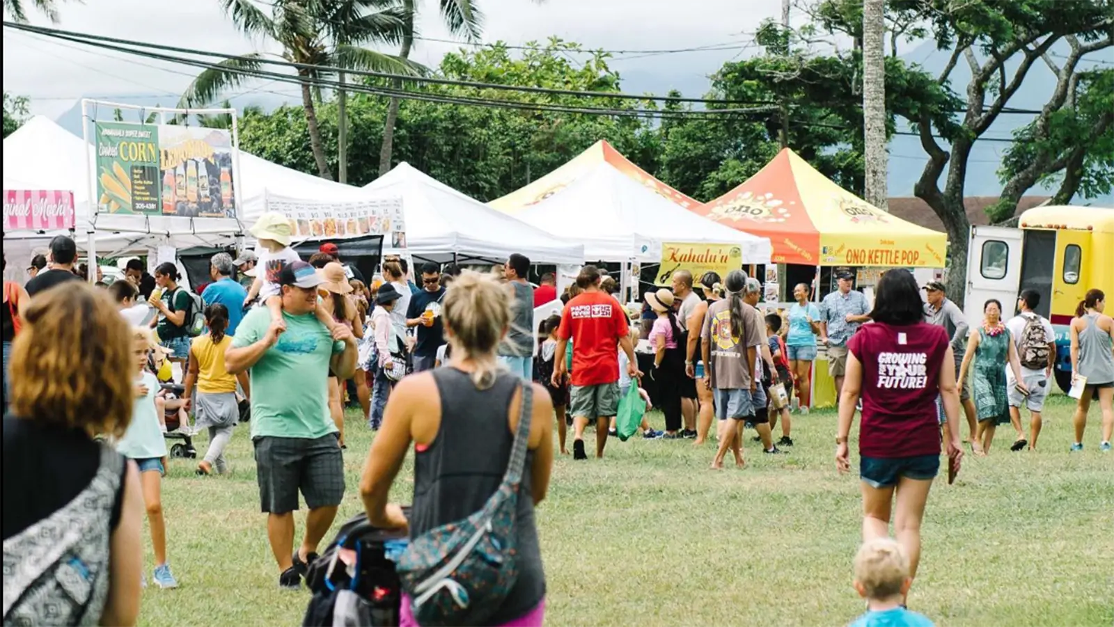 People socialize and browse vendor stalls at an outdoor festival. Various tents offer food and goods, with a shaved ice truck on the right. Lush trees and mountains are visible in the background, creating a vibrant and lively atmosphere.