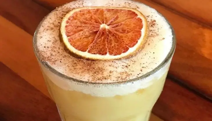 A creamy beverage in a glass, topped with frothy foam, a dusting of cinnamon, and a slice of dried orange. The glass is placed on a wooden surface, making it the best drink to enjoy during Happy Hour on Kauai's North Shore.